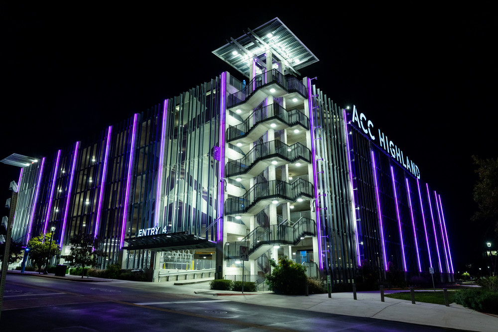 A modern multi-story parking garage illuminated at night, featuring a sleek design with vertical glass panels. The building is adorned with vibrant purple lighting, highlighting its architectural features. A prominent sign reads